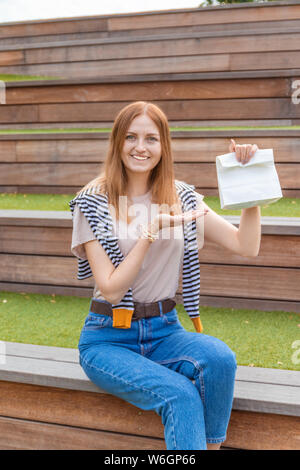 Lächelnde blonde Schüler Mädchen in T-Shirt und Blue Jeans sitzt auf einer Holzbank mit einer Papiertüte an einem sonnigen Tag in den Park. Mittagspause. Leer Stockfoto