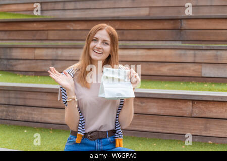 Lächelnde blonde Schüler Mädchen in T-Shirt und Blue Jeans sitzt auf einer Holzbank mit einer Papiertüte an einem sonnigen Tag in den Park. Mittagspause. Leer Stockfoto