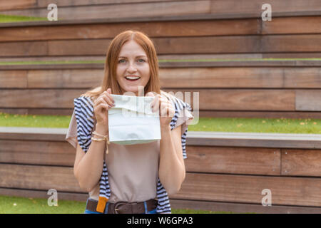 Lächelnde blonde Schüler Mädchen in T-Shirt und Blue Jeans sitzt auf einer Holzbank mit einer Papiertüte an einem sonnigen Tag in den Park. Mittagspause. Leer Stockfoto