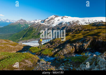 Ein Mann mit dem Rucksack in der Nähe von einem kleinen Bach in Kenai Fjords National Park mit Exit Gletscher im Hintergrund an einem sonnigen Sommertag im Süden - zentrales Alaska Stockfoto