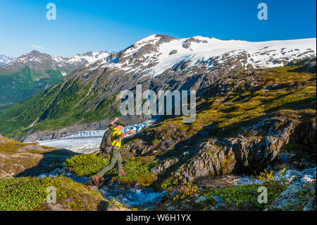 Ein Mann mit dem Rucksack in der Nähe von einem kleinen Bach in Kenai Fjords National Park mit Exit Gletscher im Hintergrund an einem sonnigen Sommertag im Süden - zentrales Alaska Stockfoto