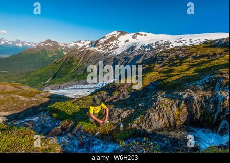 Ein Mann, der mit dem Rucksack unterwegs ist und seine Wasserflasche aus einem kleinen Bach im Kenai Fjords National Park mit dem Exit Glacier im Hintergrund auf einem sonnigen... Stockfoto