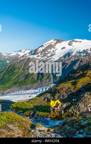 Ein Mann, der mit dem Rucksack unterwegs ist und seine Wasserflasche aus einem kleinen Bach im Kenai Fjords National Park mit dem Exit Glacier im Hintergrund auf einem sonnigen... Stockfoto