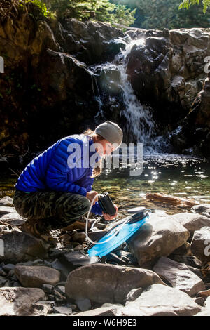 Weibliche Backpacker filterndes Wasser, beleuchtet von der Morgensonne auf dem High Divide Trail, mit Wasserfall im Hintergrund, Olympic National Park, Olymp... Stockfoto