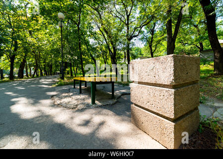 Eine gelbe Holzbank im Park im Sommer abends in der Sonne. In der Nähe befindet sich ein Quadrat konkrete Mülleimer. Stockfoto