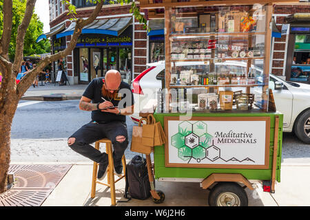 ASHEVILLE, NC, USA-27 Juli 19: eine tätowierte Mann sitzt auf einem Hocker in der Innenstadt von Asheville verkauft Meditative Heilmittel. Stockfoto