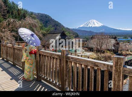 Japanische Frau im Kimono mit Papier Regenschirm im Freilichtmuseum Iyashinosato, alten japanischen Dorf mit traditionellen strohgedeckten Häuser, Vulkan zurück Stockfoto