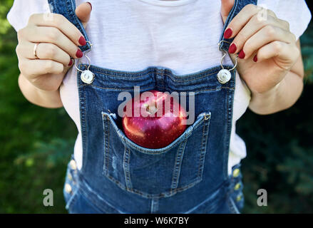 Rot reifen Apfel in die Tasche Blue Jeans Overalls. Landwirtschaft, Gartenbau, Ernte Konzept. Stockfoto