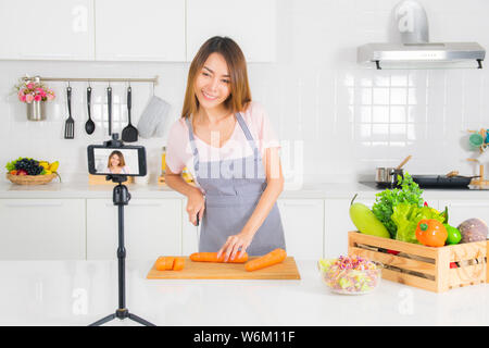 Asiatische schöne Frau, lange Haare, trägt ein rosa t-shirt und gestreifte Schürze stehend, schneiden Karotten auf dem Tisch moderne weiße Küche und Recordi Stockfoto