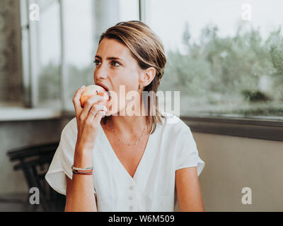 Junge Frau isst einen Apfel beissen vor dem großen Fenster der Küche ihres Hauses. Stockfoto