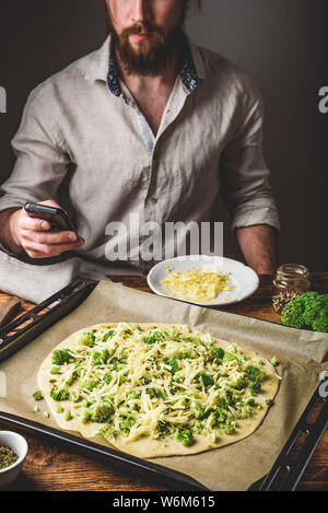 Bärtiger Mann surfen Handy beim Kochen Pizza mit Brokkoli, Pesto, Gewürze und Käse Stockfoto