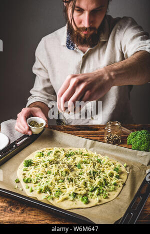 Bärtiger Mann gießt Gewürze auf rohe Pizza mit Brokkoli, Pesto und Käse Stockfoto