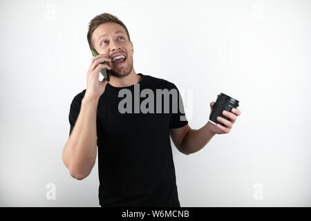 Hübscher junger Mann mit schwarzen T-Shirt sprechen auf Smartphone und Holding Tasse Kaffee sieht auf isolierten weißen Hintergrund zufrieden Stockfoto