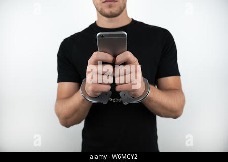 Mann, schwarzes T-Shirt, Smartphone, während er mit Handschellen gefesselt auf dem isolierten weißen Hintergrund Stockfoto
