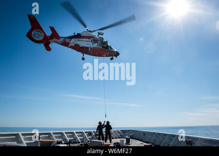 Mitglieder der Besatzung an Bord der Coast Guard Cutter Stratton verhalten Korb heben die Entwicklungen, die mit dem Air Crew an Bord eines MH-65 Dolphin Helikopter in die Coral Sea Juli 12, 2019. Der Korb abgesenkt wird und wieder nach oben gehoben, damit die Crew in realistischen Szenarien zu trainieren und ihre Kenntnisse sowohl bei den Besatzungen zu errichten. (U.S. Coast Guard Foto von Petty Officer 2. Klasse Jasmin Mieszala) Stockfoto