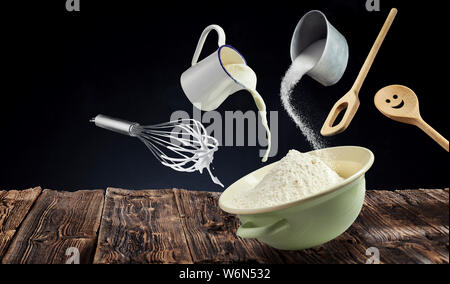 Composite freeze motion Konzept, Teig mit Mehl in eine Schüssel unter abgehängten Holz- Utensilien, Schneebesen, Milch gießt aus einem Krug und Mehl sprinkl Stockfoto