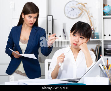 Porträt der jungen betonte asiatischen Geschäftsfrau in Multirassischen Office mit den Unzufriedenen weiblichen chief Schelte hinter Stockfoto