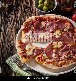 In Scheiben geschnittene Peperoni oder Salami Pizza gewürzt mit Kräutern und mit einer Schüssel mit Oliven auf rustikalen, mit Holz im quadratischen Format serviert. Stockfoto