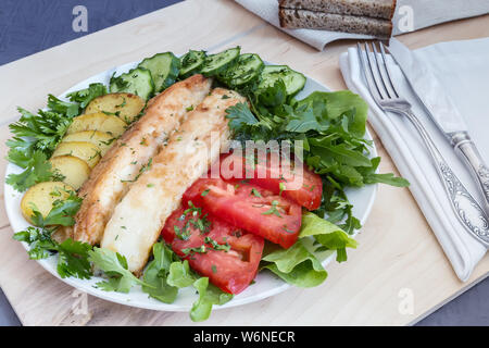 Auf dem Tisch auf einem Teller zwei Scheiben gebratener Fisch, gebratene Kartoffeln, Gurken, Tomaten, Salat, Petersilie und Dill. Stockfoto