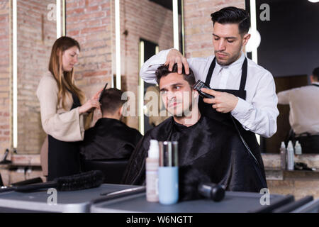 Professionelle friseur Mann tun Styling hübscher Kerl mit elektrischer Haarschneider bei Friseur Stockfoto