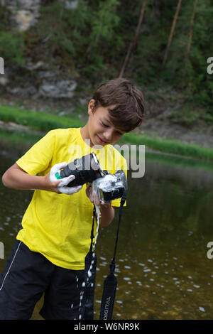 Ein 9-jähriger Junge im Wasser hält eine SOAP-Kamera und einem Schwamm in seinen Händen. falsche Reinigung der Kamera und des Objektivs Stockfoto