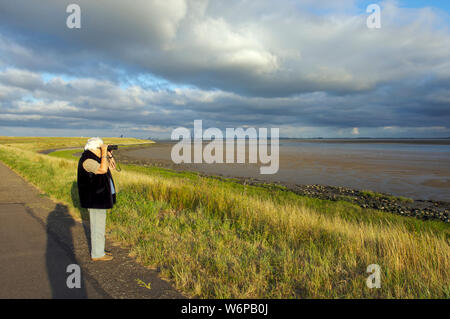 Active Senior mit Fernglas mit Blick auf die Westerschelde bei Ebbe mit Doel Kernkraftwerk im Hintergrund in Zeeland, Niederlande Stockfoto