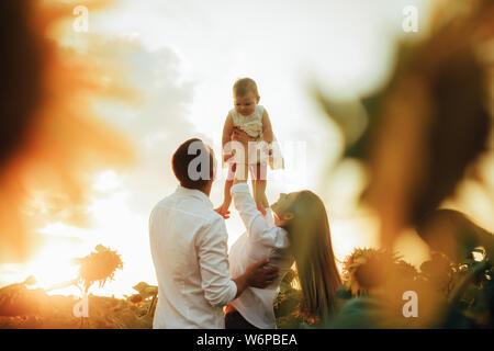 Glückliche junge Familie mit Baby hat einen Spaß und spielt im Sonnenblumenfeld bei Sonnenuntergang. Stockfoto