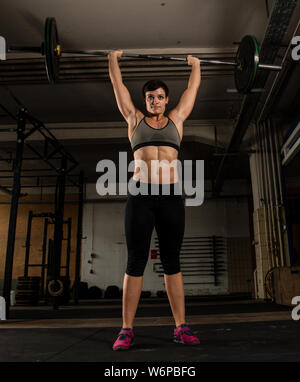 Eine sportliche kurze behaarte Frau mit starken abs tut Gewichtheben mit einem barbell in ein Fitnessstudio. Die muskulöse Frau tut eine Schulter, an der Oberleitung reinigen. Stockfoto