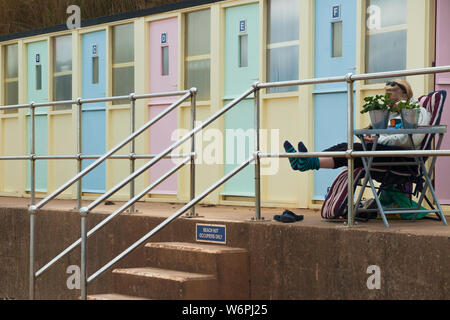 Frau/Person sitzt Lesen/in einem Liegestuhl/Liegestuhl vor der Küste Meer Strand sitzen Hütten/Seaside Beach Hut in Sidmouth Devon, Großbritannien. (110) Stockfoto