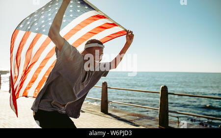 Lächelnd afrikanischer Mann an der Strandpromenade mit einer amerikanischen Flagge. Fröhlicher junger Kerl an der Promenade, die amerikanische Flagge. Stockfoto