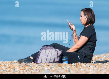 Seitliche Sicht auf eine junge Frau sitzt auf einem Strand können Sie über Ihr Smartphone mit Kopfhörer Musik zu hören. Über ein Telefon am Strand. Stockfoto