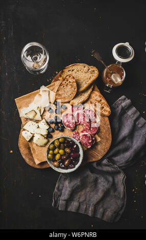 Wein und Snacks. Flachbild-lay gegrilltes Stück Brot, Käse, Salami, Oliven, Aprikosenmarmelade, Beeren und weißen Wein über rustikale Schwarz Stockfoto