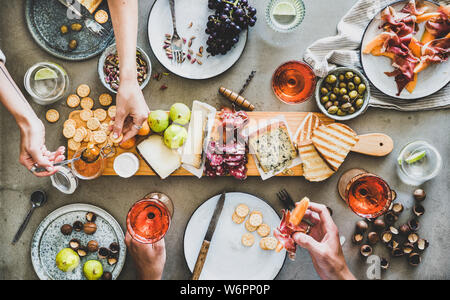 Mitte Sommer Picknick mit Wein und Snacks. Flachbild-lay von Wurst- und Käseplatte, Rose Wein, Nüsse, Oliven und Völker Hände mit Snacks auf Beton Stockfoto