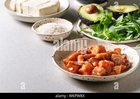 Zutaten zum Kochen poke Schüssel. Reis, Soja Sauce marinierter Lachs, Avocado, Tofu Käse und grünem Salat in Keramik Schalen auf Grau gefleckt. Stockfoto