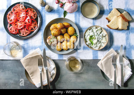 Sommer vegetarisch Essen für die Familie oder Freunde. Junge gebackene Kartoffeln, Tomaten Carpaccio, Gurken Salat, Brot, Wein, Soße und Blumen über Leitung Stockfoto