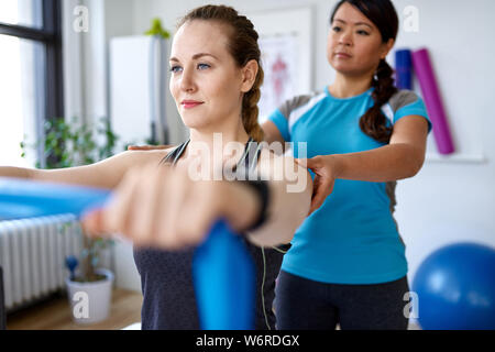 Chinesische Frau Physiotherapie Professional eine Behandlung durch die Verwendung elastischer Widerstand Bänder zu einer attraktiven Blondine Client in einem hellen Medizinisches Büro Stockfoto