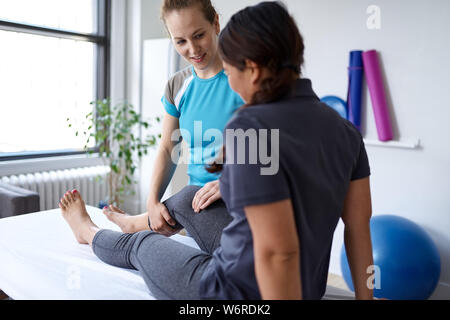 Kaukasische Frau Physiotherapeutin strectching das Bein und Knie eines mittleren nach chinesischen weiblichen Patienten sitzen auf einem Massagetisch Stockfoto