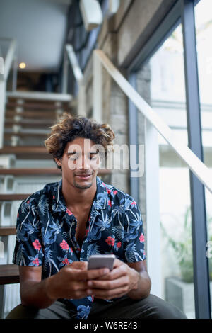 Attraktive ethnischen Hipster tausendjährigen business Mann lächelnd und Lesen blog Handy sitzen auf moderne Treppe der helle Zimmer mit tropischen Pflanzen Stockfoto