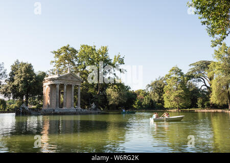 Horizontale Ansicht des Aesculapius Tempel in der Villa Borghese durch Vegetation, an einem sonnigen Tag umgeben. Boote mit Touristen Schwimmen im Teich. Stockfoto