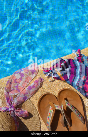 Sommer flatlay, Stroh Hut mit Schal, Strand wickeln und Sandalen an der Seite eines blau gefliesten Pool. mit Platz kopieren Stockfoto