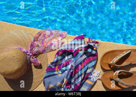 Sommer flatlay, Stroh Hut mit Schal, Strand wickeln und Sandalen an der Seite eines blau gefliesten Pool. mit Platz kopieren Stockfoto