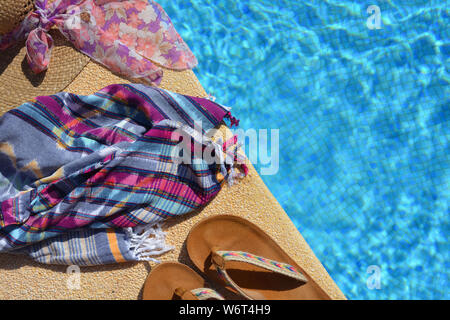 Sommer flatlay, Stroh Hut mit Schal, Strand wickeln und Sandalen an der Seite eines blau gefliesten Pool. mit Platz kopieren Stockfoto