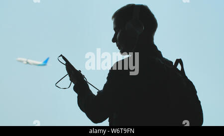 Ein junger Kerl dreht sich auf Musik auf einem Smartphone und setzt auf Kopfhörer vor dem Hintergrund der ein Flugzeug. Stockfoto