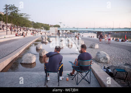 LYON, Frankreich - Juli 18, 2019: Zwei Männer sitzen am Ufer der Rhone (QUAIS) nach Pont de la Guillotiere Brücke am Abend, während die Menschen ein Stockfoto