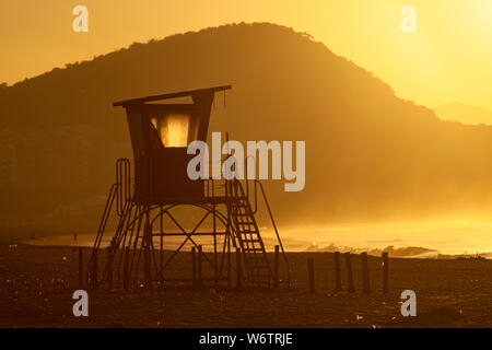 Silhouette eines alten Rettungsschwimmer Turm vor einem leuchtenden Dunst am Strand von Copacabana in Rio de Janeiro mit Hintergrundbeleuchtung durch die intensiven goldenen Stunde Sonnenlicht mit Stockfoto