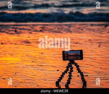 Sonnenuntergang über der Küste. Strandwellen im Hintergrund. Smartphone-Kamera auf einem Stativ im Vordergrund Stockfoto