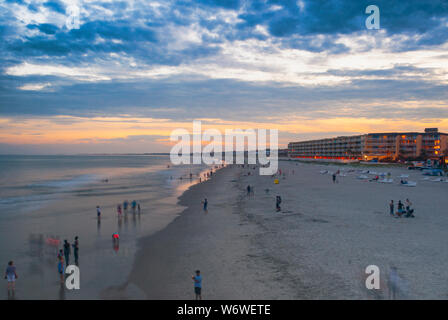 Die Sonne geht an Folly Beach, South Carolina, während die Leute am Wasser entlang laufen. Es gibt einige Leute spielen im Sand und ein Hotel auf der rechten Seite. Stockfoto