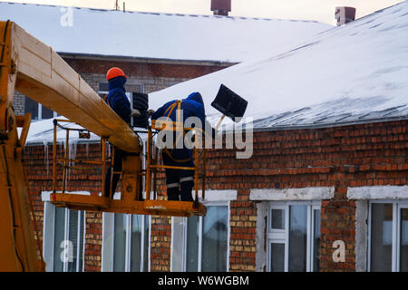 Arbeitnehmer in orangefarbenen Overalls und Helme auf dem Kran Warenkorb Eiszapfen vom Dach des Hauses nehmen Sie an einem Wintertag - Reinigung der Überdachung, utility Servi Stockfoto