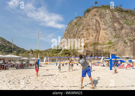 Jugendliche serviert in einer Partie Beachvolleyball am Copacabana Strand an einem sonnigen Tag mit dem Leme Berg im Hintergrund Stockfoto