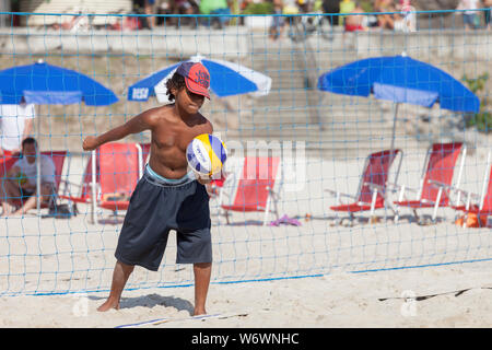 Nahaufnahme von konzentrierter Jugendlicher, kurz bevor er serviert in einer Partie Beachvolleyball mit Sonnenliegen und Schirmen im Hintergrund Stockfoto